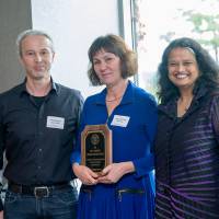 A color photo showing a man and two women posing for a photo. The woman in the middle is holding an award