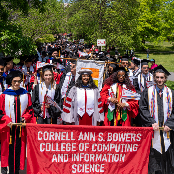 Several students in graduation robes march down a street on sunny day holding a banner that says Cornell Ann S. Bowers College of Computing and Information Science.