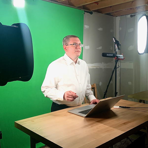 Color photo of a man in a white dress shirt and glasses being recorded in a studio