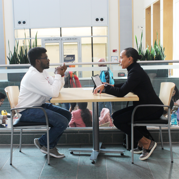A color photo of two people sitting at a table, talking to each other
